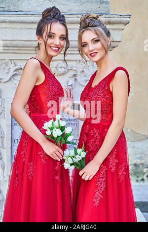 Two beautiful bridesmaids girls blonde and brunette ladies wearing elegant full length red chiffon bridesmaid dress with lace and holding flower Stock Photo