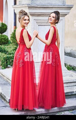 Two beautiful bridesmaids girls blonde and brunette ladies wearing elegant full length red chiffon bridesmaid dress with lace and holding flower Stock Photo