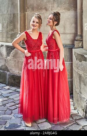 Two beautiful bridesmaids girls blonde and brunette ladies wearing elegant full length red chiffon bridesmaid dress with lace and holding flower Stock Photo