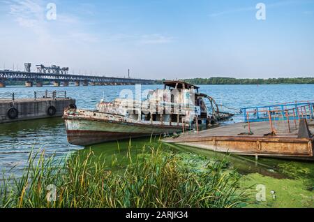 Semi-flooded river pleasure boat at an abandoned pier on background of railway bridge Stock Photo