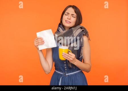 Cold treatment. Unhappy sick woman wrapped in scarf standing with closed eyes, going to sneeze, holding tissue and cup of tea, feeling unwell with inf Stock Photo