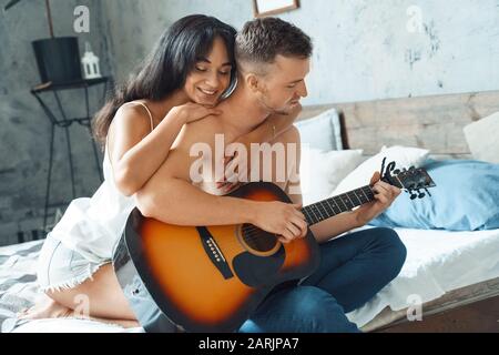 Mixed Race Couple. Young man playing guitar while woman hugging him cheerful sitting on bed at home Stock Photo