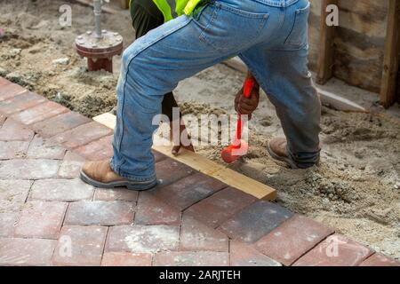 Brick layer tamping or tapping his orange mallet or hammer against a 2x4 to adjust the freshly laid pavers on the walkway. Stock Photo