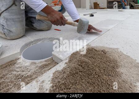 paver tapping or tamping on a granite concrete tile and setting it in place after cutting a curve out if it to fit around a drain pipe. Construction Stock Photo