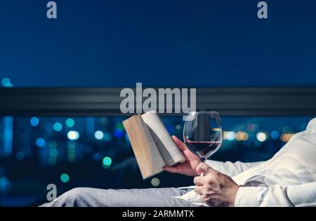 The man relaxing in the room reading book with a glass of red wine and background of city bokeh lights. Stock Photo