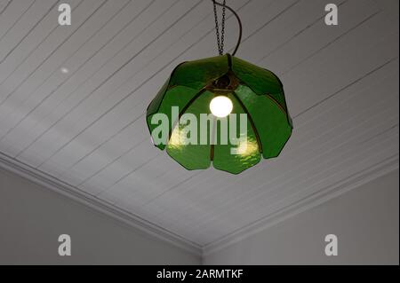 A room is lit up by an electric light that has an old fashioned, heavy green glass lampshade hanging from the ceiling Stock Photo