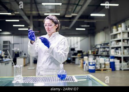 Serious lab woman in white coat with experimental glass in her hands conducts experiments on defocused background Stock Photo