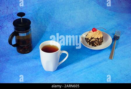 Tea break, white ceramic tea mug, teapot and saucer with biscuit cake in chocolate chips and cherries. Close-up. Stock Photo