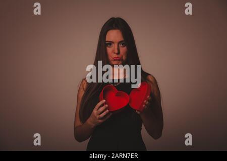 Valentine day. Sad upset grymacing woman in black opening empty heart box gift present. Elegant attractive model with valentines symbol sign. Studio s Stock Photo
