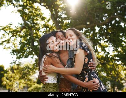 Portrait of happy three multiethnic multiracial female friends closely hugging and showing care and love for each other in park Stock Photo