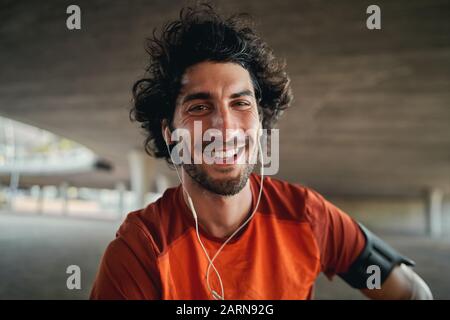 Portrait of happy young male runner with earphones in his ears looking at camera outdoor smiling - excited fit male
