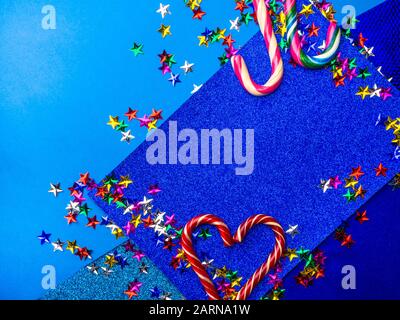Happy holidays greeting card with striped candy canes in a heart shape with a colorful stars on trendy blue background with copy space. Birthday Stock Photo