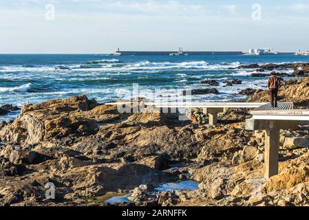 Woman walks on a wooden footpath above the rocks of Atlabtic Ocean shore in Nevogilde civil Parish of Porto city in Portugal Stock Photo