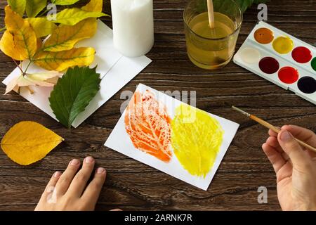 The child draws a candle and watercolor paints autumn leaves on wooden background. Crafts from paper for children. Children's art project. Stock Photo