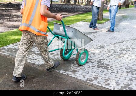 Professional gardener worker walkink with wheel-barow at park construction site. process of installation grass roll carpet and paving sidewalk on Stock Photo