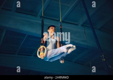 Little male gymnast training in gym, flexible and active. Caucasian fit boy, athlete in white sportswear practicing in exercises for balance on the rings. Movement, action, motion, dynamic concept. Stock Photo
