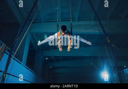 Little male gymnast training in gym, flexible and active. Caucasian fit boy, athlete in white sportswear practicing in exercises for balance on the rings. Movement, action, motion, dynamic concept. Stock Photo