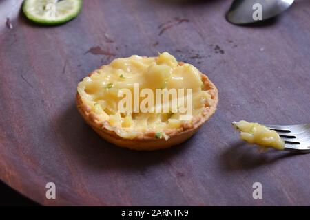 half eaten lemon tart with fork on a wooden surface with  green sliced lemon in the background Stock Photo
