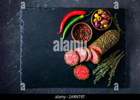 salami coated with whole green peppercorns and have sliced on a black stone board and served with olives, thyme sprigs and peppers, flat lay, close-up Stock Photo
