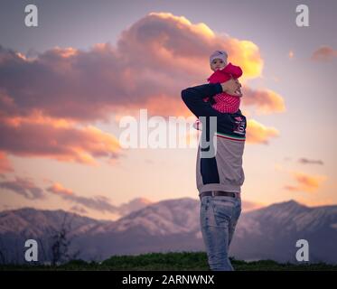 portrait of a baby girl riding on top of her father's shoulders at mountains during sunset with beautiful cloudscape in the background. Stock Photo