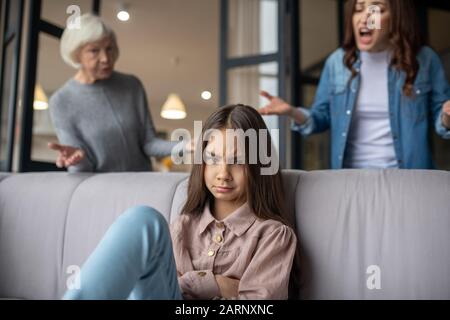 Upset daughter sitting on the couch, mom cursing. Stock Photo