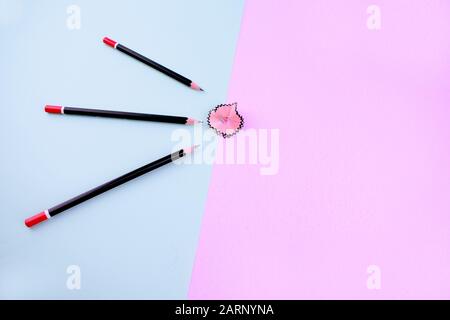 three dark black sharpened pencils over a light blue background are pointing at a pink pencil shaving which on a pink paper background Stock Photo