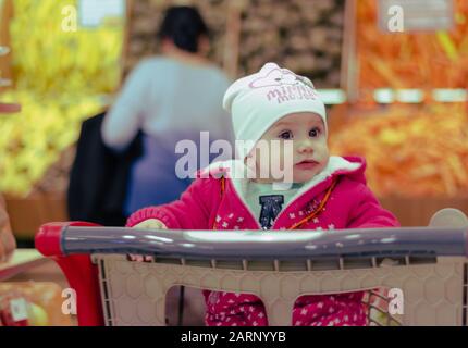 a cute little girl sitting and waiting in the shopping trolley while her grandmother buying vegetables in the grocery shop. Stock Photo