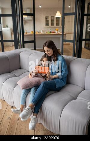 Daughter with tablet in her hands and mom sitting on couch. Stock Photo