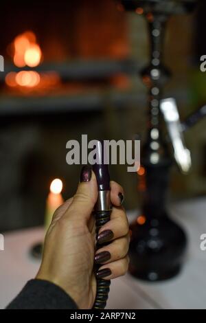 Hookah mouthpiece with a stream of smoke in a beautiful female hand on a dark background. hand and hookah pipe. Hookah in the dark at night against th Stock Photo