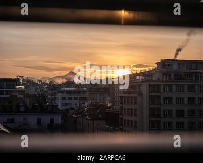 Taken through the blinds of an office window. A Sunrise over city buildings and rooftops. The sun appears behind a building with mountains in the back Stock Photo