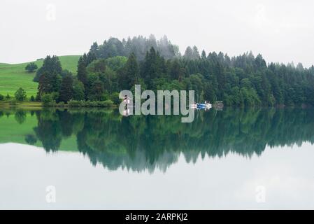 Boathouse and boats on a grove at the shore at Forggensee refelcting in the water  on a foggy overcast day, Allgäu, Bavaria, Germany Stock Photo
