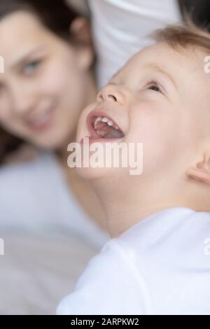 Croon-plan toddler in white looks up and smiles in front of mother Stock Photo