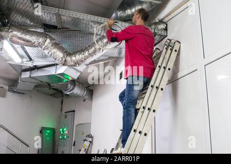 A man standing on the stairs is repairing the ventilation ducts. Maintenance of communications is performed in the technical room. The installer works Stock Photo