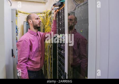 The system administrator works in the server room. The network engineer switches the wires in the data center. A young man is standing next to the rac Stock Photo