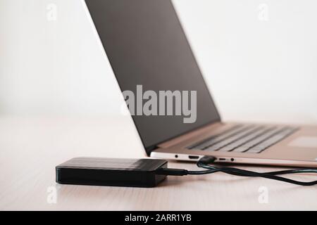 Black external hard disk, hdd on wooden table connected with mini usb cable to laptop, white background. Color image, modern technology concept. Stock Photo