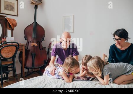 Three little sisters indoor at home using tablet lying in bed supervised by parents - education, security, streaming concept Stock Photo