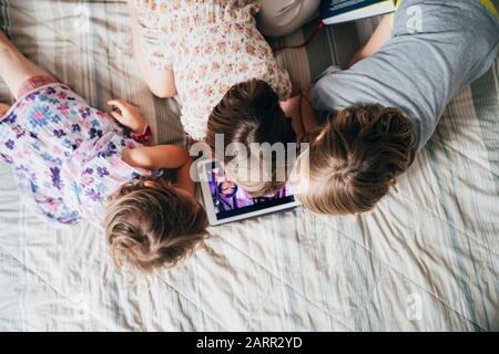 Overhead view of three little sisters indoor at home using tablet lying in bed - education, technology, streaming concept Stock Photo