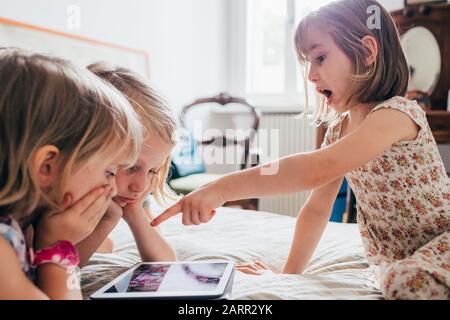 Three little sisters indoor at home using tablet lying in bed - education, technology, streaming concept Stock Photo