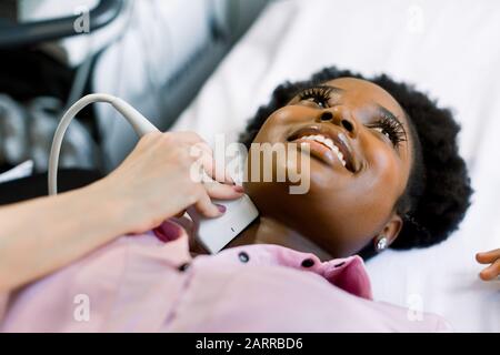 Close up shot of a young smiling African American woman getting her neck and thyroid gland examined by doctor using ultrasound scanner at the modern Stock Photo