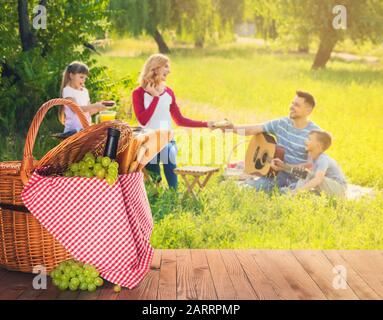 Wicker basket with tasty food and drink on table near happy family on summer picnic Stock Photo