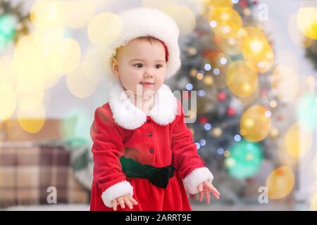 Cute little baby in Santa Claus costume and blurred lights in room decorated for Christmas Stock Photo