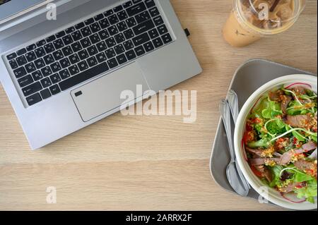 Flat lay, Modern laptop with spicy pork salad and ice coffee on wooden table Stock Photo