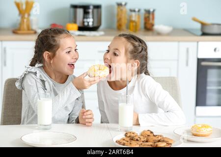 Portrait of cute twin girls eating donuts with milk in kitchen Stock Photo