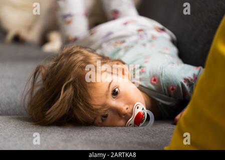 Cute 2 year old Baby girl on a bed on her belly with head on sofa. Bright interior. Stock Photo