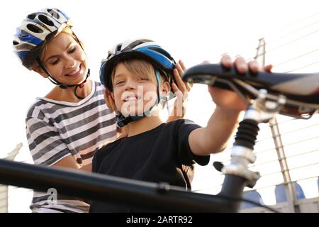 Mother helping her son to put on bicycle helmet outdoors Stock Photo