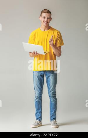 Young man in stylish t-shirt and with laptop showing thumb-up gesture on grey background Stock Photo