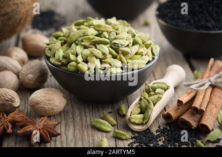 Green cardamom pods in black ceramic bowl. Aromatic spices: anise, gloves, black cumin seeds, nutmegs, cinnamon sticks, turmeric. Ingredients for heal Stock Photo