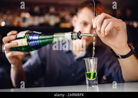 Bartender is preparing mix of cocktail with spoon of green alcohol in small glass, bar counter Stock Photo