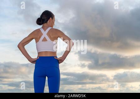 Back view of a sporty woman with her hands on her hips, resting after exercising, sunrise, sunset. Stock Photo