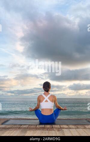 woman practicing yoga, sitting in Easy Seat exercise, Sukhasana pose, working out, meditation session. Back view with ocean background Stock Photo